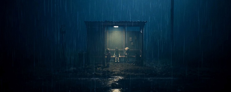 Dos figuras de pie bajo la lluvia junto a un cobertizo iluminado